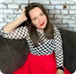Young woman with brown hair wearing a black and white checkered top and red skirt sitting in front of a white brick wall.